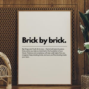 以下が含まれることがあります： 「Brick by brick」という黒い文字が書かれた額入りのプリント。プリントは白い背景で、木の壁に配置されています。フレームは白です。メインタイトルの下にも追加のテキストが含まれています。