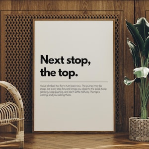 May include: A framed print with the text "Next stop, the top." in a black, bold font. The print is set against a wooden wall with a woven chair and a potted plant in the background.