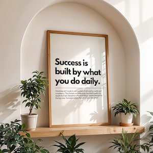 May include: A framed print with the text "Success is built by what you do daily." The print is in a light wood frame and sits on a wooden shelf. Two potted plants flank the print, with more greenery in the foreground.