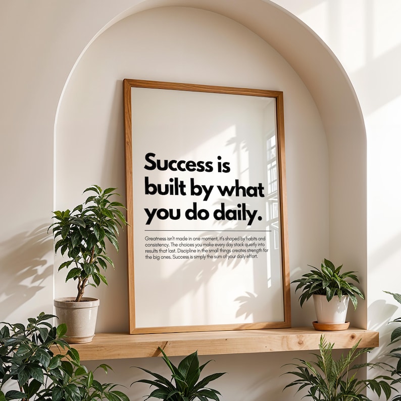 May include: A framed print with the text "Success is built by what you do daily." The print is in a light wood frame and sits on a wooden shelf. Two potted plants flank the print, with more greenery in the foreground.