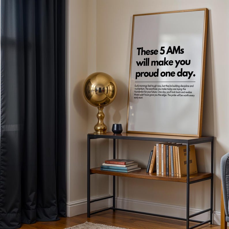 Pu&ograve; includere: Una stampa incorniciata con il testo "These 5 AMs will make you proud one day" poggia su una mensola in metallo nero e legno. Una lampada a sfera dorata e libri sono anche sulla mensola. Una tenda scura &egrave; a sinistra.