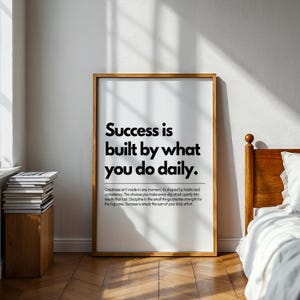May include: A framed print with the text "Success is built by what you do daily." The print has a wooden frame and is displayed in a room with a bed and a stack of books. The text is in black.