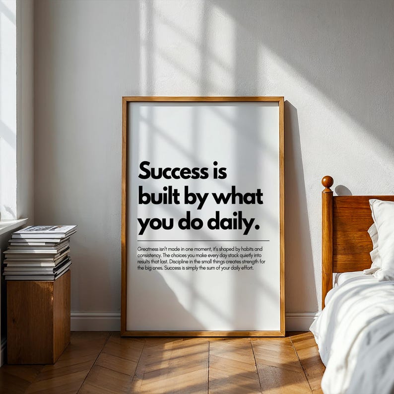 May include: A framed print with the text "Success is built by what you do daily." The print has a wooden frame and is displayed in a room with a bed and a stack of books. The text is in black.