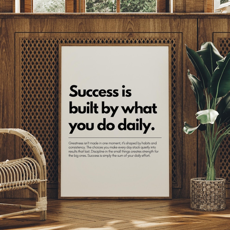 May include: Framed print with the text "Success is built by what you do daily." The print is in a white frame, set against a wooden wall. A potted plant and a wicker chair are also visible.