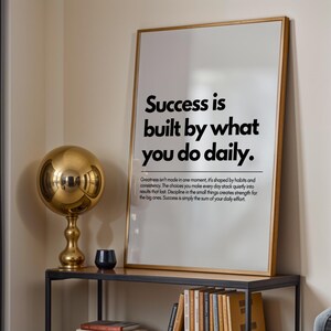 May include: A framed print with the text "Success is built by what you do daily." A gold-colored sphere lamp and a small black candle holder sit on a black metal shelf. Books are visible below the shelf.