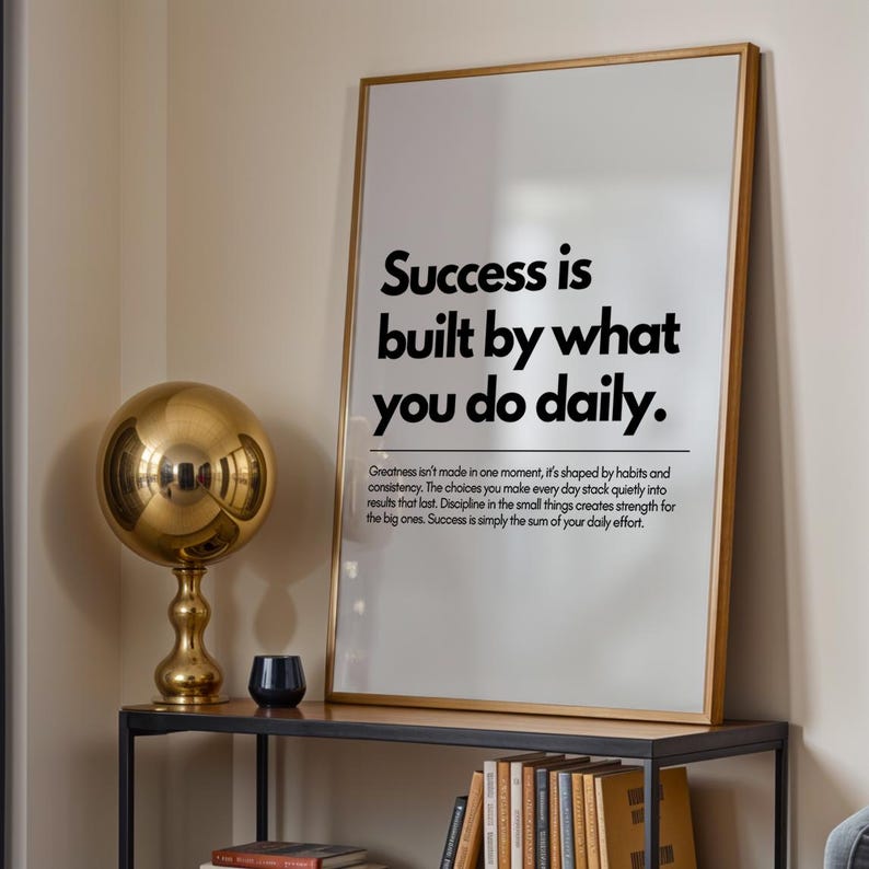 May include: A framed print with the text "Success is built by what you do daily." A gold-colored sphere lamp and a small black candle holder sit on a black metal shelf. Books are visible below the shelf.