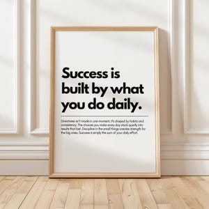 May include: A framed print with the text "Success is built by what you do daily." The print is in a light wood frame and is displayed against a white wall. The text is in a bold, black font.