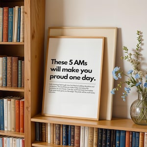 Pu&ograve; includere: Una stampa incorniciata con il testo "These 5 AMs will make you proud one day." La stampa &egrave; in una cornice di legno e poggia su una mensola di legno con una libreria sullo sfondo. Un vaso di fiori blu &egrave; a destra.