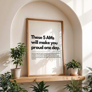 Pu&ograve; includere: Una stampa incorniciata con il testo "These 5 AMs will make you proud one day." La stampa &egrave; in una cornice di legno chiaro e si trova su una mensola di legno. Piante in vaso sono posizionate intorno alla stampa.