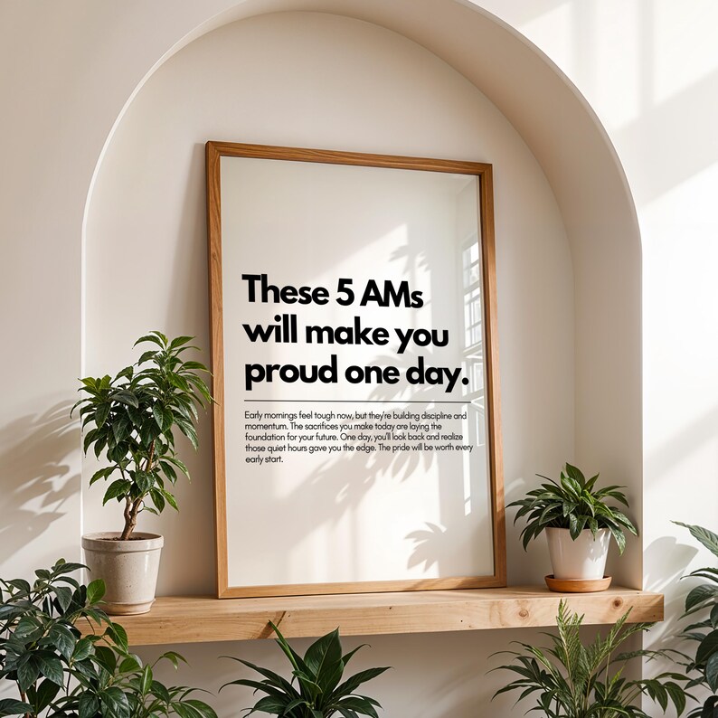 Pu&ograve; includere: Una stampa incorniciata con il testo "These 5 AMs will make you proud one day." La stampa &egrave; in una cornice di legno chiaro e si trova su una mensola di legno. Piante in vaso sono posizionate intorno alla stampa.