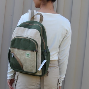 May include: A green, beige, and brown hemp backpack. The backpack features a main compartment, a front pocket, and side pockets. It has a tag attached. The backpack displays the text "Himalayan" and a cannabis leaf logo.