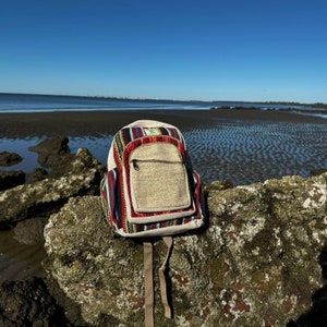 Puede incluir: Una mochila de cáñamo multicolor con un gran bolsillo frontal. La mochila está sentada en una roca con una playa y el océano en el fondo.