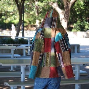 May include: A patchwork jacket with a hood, featuring a variety of colors and patterns. The jacket is made of different fabric squares in shades of red, green, blue, and brown. The person is wearing blue jeans.