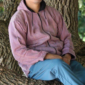 May include: A person wearing a pink and black zip-up hoodie and blue pants, sitting on a tree branch. The hoodie has a textured pattern. The person is wearing sunglasses.