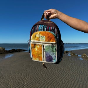 May include: A multi-coloured woven backpack with a brown leather strap and a green leaf logo. The backpack has a front pocket with a zipper and a main compartment with a flap closure.