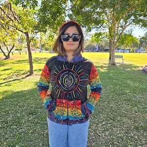 May include: A black hooded jacket with a colourful embroidered sun design. The jacket has a zip closure and pockets on the front. The sleeves are long and the jacket is made of a soft, comfortable fabric.