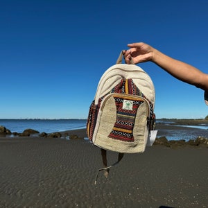 Pode incluir: Uma mochila de cânhamo bege com um padrão tecido colorido na aba frontal. A mochila tem um pequeno logotipo de folha verde com o texto "Hemptique" nela.