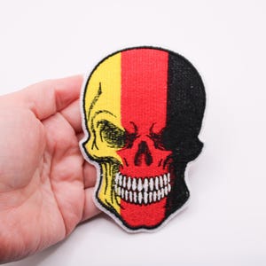 Op de afbeelding: Geborduurde schedelpatch met een ontwerp met gele, rode en zwarte verticale strepen, die op een vlag lijken. De schedel heeft een gedetailleerde, open mond met witte tanden. Witte achtergrond.