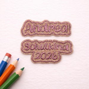 Könnte beinhalten: Zwei gestickte Aufnäher mit dem Namen "Andrea" und dem Text "Schulkind 2026" in Schreibschrift. Die Aufnäher haben einen glitzernden Goldhintergrund mit rosa Rand und Schrift. Drei Buntstifte befinden sich in der linken unteren Ecke.