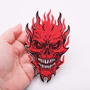 Op de afbeelding: Geborduurde patch met een rode schedel in vlammen. De schedel heeft een dreigende uitdrukking met scherpe tanden en zwarte details. De vlammen zijn zwart omlijnd, wat een sterk contrast creëert met het rood.
