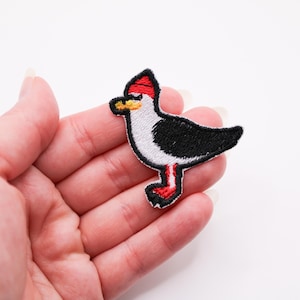 Puede incluir: Parche bordado de un pájaro de dibujos animados con cresta roja, alas negras y patas rojas. El pájaro tiene un cuerpo blanco y un pico amarillo. El parche está delineado en negro y se sostiene en una mano.