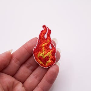 Può includere: Toppa ricamata a forma di fiamma. Presenta una vivace combinazione di colori rosso, arancione e giallo, con dettagli bianchi. Il design della fiamma è dettagliato con cuciture intricate, creando un aspetto strutturato.