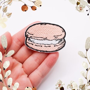 Może przedstawiać: Naszywka z haftowanym różowym i białym makaronikiem. Makaronik to popularne francuskie ciastko.