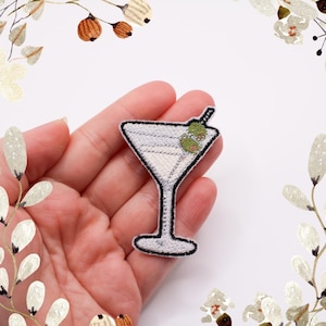 Könnte beinhalten: Gestickter Aufnäher mit einem Martini-Glas mit Oliven. Der Aufnäher hat einen weißen Hintergrund und eine schwarze Umrandung. Die Oliven sind grün. Der Aufnäher ist für Kleidung oder Accessoires geeignet.