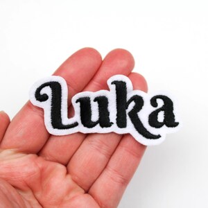 Peut inclure: Un écusson blanc avec le nom "Luka" brodé en lettres cursives noires. L'écusson a une bordure noire et est tenu dans la main d'une personne.