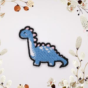 Puede incluir: Parche azul bordado de un dinosaurio de dibujos animados con detalles rosas.