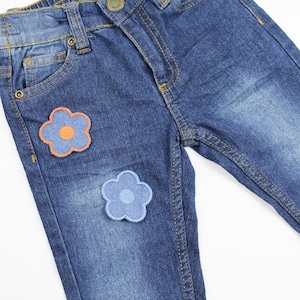 Pode incluir: Jeans azul denim com dois patches bordados em forma de flor, um laranja e outro azul.