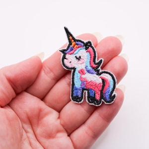 May include: Embroidered unicorn patch with a black outline. The unicorn has a black horn, pink, blue, and purple mane and tail, and a pink bow. The patch is held in a hand against a white background.