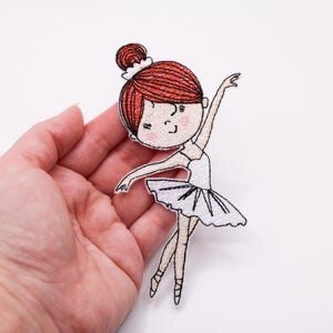 Può includere: Toppa ricamata di una ballerina con i capelli rossi raccolti in uno chignon, che indossa un tutù bianco e scarpette da punta. La toppa è tenuta in mano su sfondo bianco. La ballerina è in posa aggraziata.