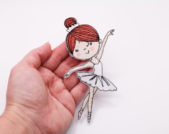 Op de afbeelding: Geborduurde patch van een ballerina met rood haar in een knot, gekleed in een witte tutu en spitsschoenen. De patch wordt in een hand gehouden tegen een witte achtergrond. De ballerina staat in een sierlijke pose.