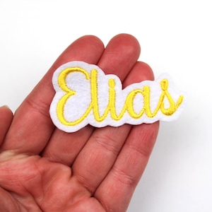 Puede incluir: Un parche de fieltro blanco con el nombre "Elias" bordado en amarillo con escritura cursiva. El parche tiene un borde blanco. El parche está en la mano de una persona.
