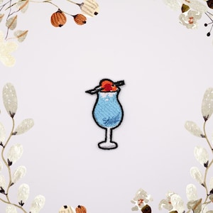 Op de afbeelding: Een geborduurde strijk-op patch van een blauwe cocktail met een rode kers erop. De drank zit in een glas met steel en een rietje.