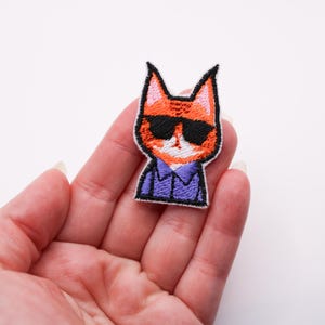 Könnte beinhalten: Gestickter Aufnäher mit einer Cartoon-Katze mit orangefarbenem Fell, schwarzer Sonnenbrille und einem lila Hemd. Die Katze hat eine weiße Schnauze und rosa Innenohren. Der Aufnäher wird in einer Hand vor weißem Hintergrund gehalten.