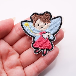May include: Embroidered patch featuring a cartoon fairy with brown hair, light blue wings, and a red dress. The fairy holds a yellow wand and a tooth. The patch has a black border and is held in a hand.