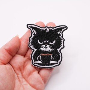 May include: Black embroidered iron-on patch of a cat holding a coffee cup. The cat has a grumpy expression.
