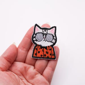 Puede incluir: Parche bordado con un gato que lleva gafas de sol y una camisa naranja con lunares negros. El gato tiene la cara blanca, pelaje a rayas grises y negras y orejas rosas. El parche se sostiene en una mano.