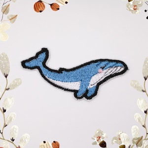 May include: Embroidered patch of a blue whale with a black outline. The whale has a white belly and a small pink blush on its cheek. The patch is designed in a cute, cartoon style, suitable for adding to clothing or accessories.