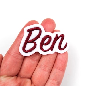 Könnte beinhalten: Ein weißer, gestickter Aufnäher mit dem Namen "Ben" in burgunderfarbener Schrift. Der Aufnäher hat einen weißen Rand und ist wahrscheinlich zum Aufnähen oder Aufbügeln auf Stoff gedacht. Der Aufnäher wird in einer Hand gehalten.