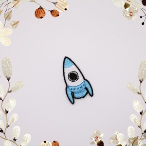 Könnte beinhalten: Ein gestickter Raketen-Patch in Blau und Weiß. Die Rakete zeigt nach oben und hat ein schwarzes Auge.
