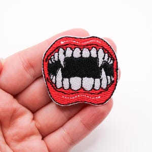 May include: Embroidered patch featuring a red mouth with sharp white teeth and a black interior. The patch is a close-up view, showcasing the detailed stitching and design. The patch is held in a hand.