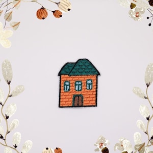 Può includere: Un piccolo ricamo a forma di casa con tetto verde e muri in mattoni arancioni. La casa ha tre finestre e una porta.