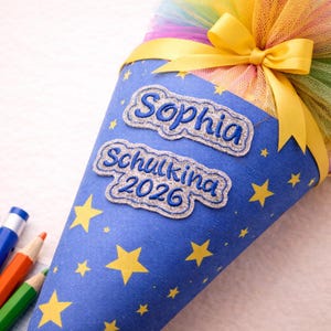 Puede incluir: Una bolsa escolar azul en forma de cono con estrellas amarillas y el nombre "Sophia" bordado. La bolsa está decorada con un lazo amarillo y tul colorido. El texto "Schulkina 2026" también está bordado en la bolsa.