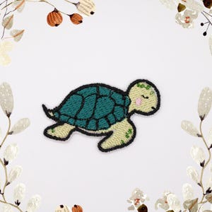 Puede incluir: Un parche bordado de una tortuga marina verde y amarilla. La tortuga tiene un caparazón verde y un cuerpo amarillo con detalles verdes. La tortuga tiene la nariz rosa y los ojos cerrados.