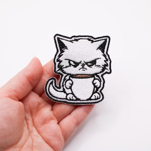 Puede incluir: Parche blanco bordado para planchar de un gato de dibujos animados enfadado con contornos negros. El gato está de pie con las patas cruzadas y tiene una expresión de enfado.