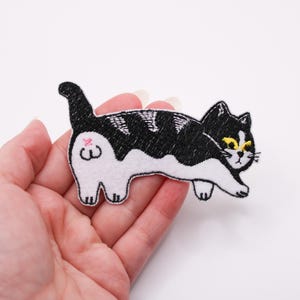 Puede incluir: Parche bordado de un gato blanco y negro con ojos amarillos y un trasero rosa. El gato está en una pose de caminar con la cola levantada. El parche se sostiene en una mano.