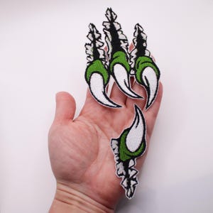 以下が含まれることがあります： 白、緑、黒の刺繍された爪パッチ。パッチは、白い爪、緑のパッド、黒のディテールを備えた爪のデザインが特徴です。衣類やアクセサリーにユニークなタッチを加えるのに最適です。